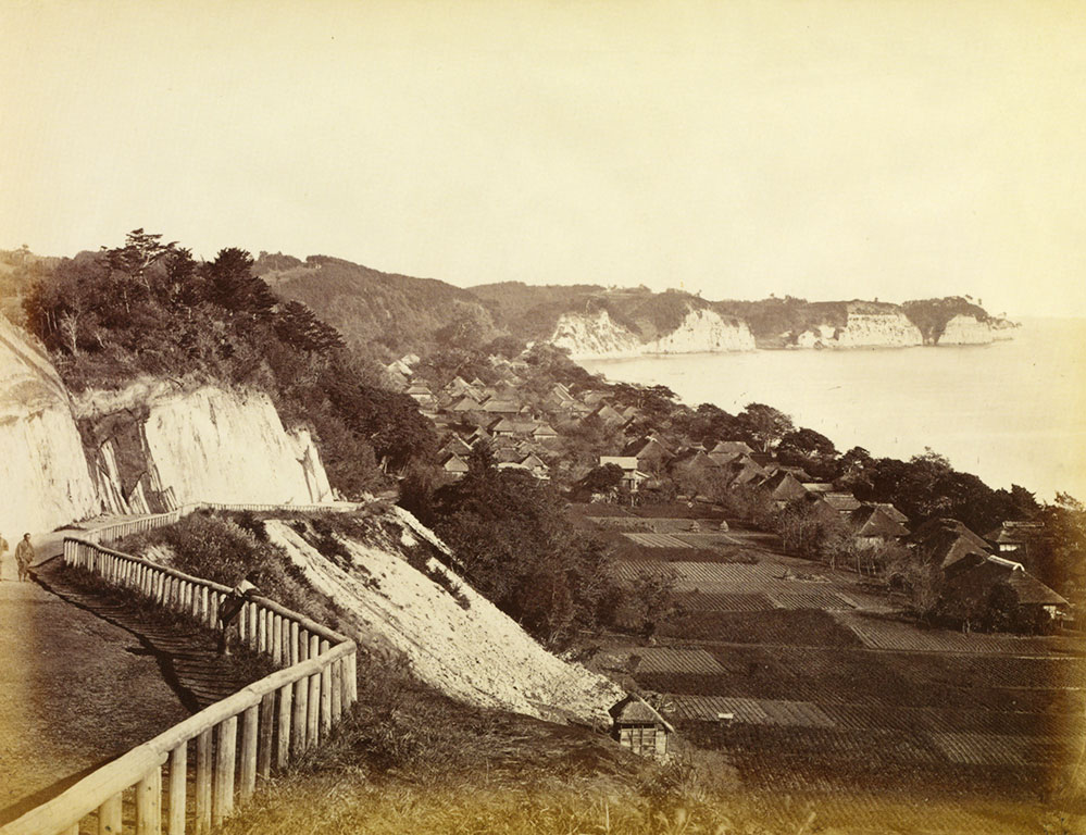 Fenced path along a cliff, fields and houses on a bay