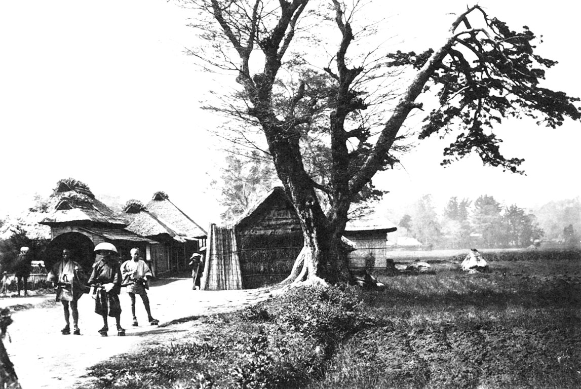 Three people and a tree in the foreground, huts in the background