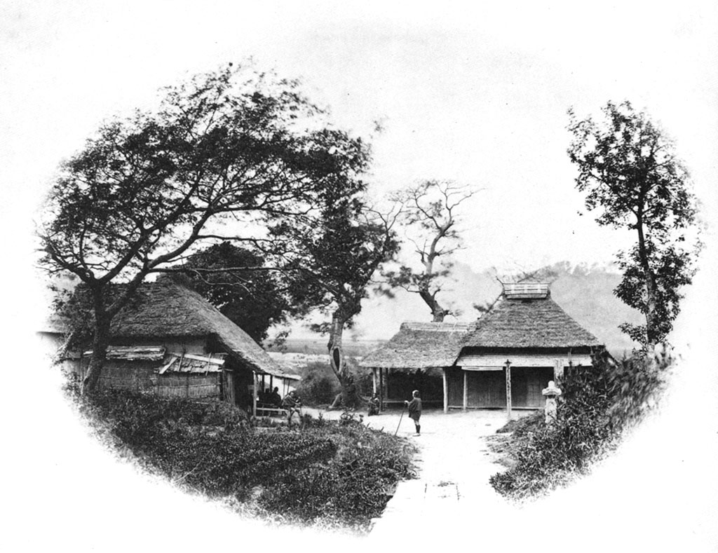 Two huts, trees in the foreground, a person standing in the center of the image, facing to the left