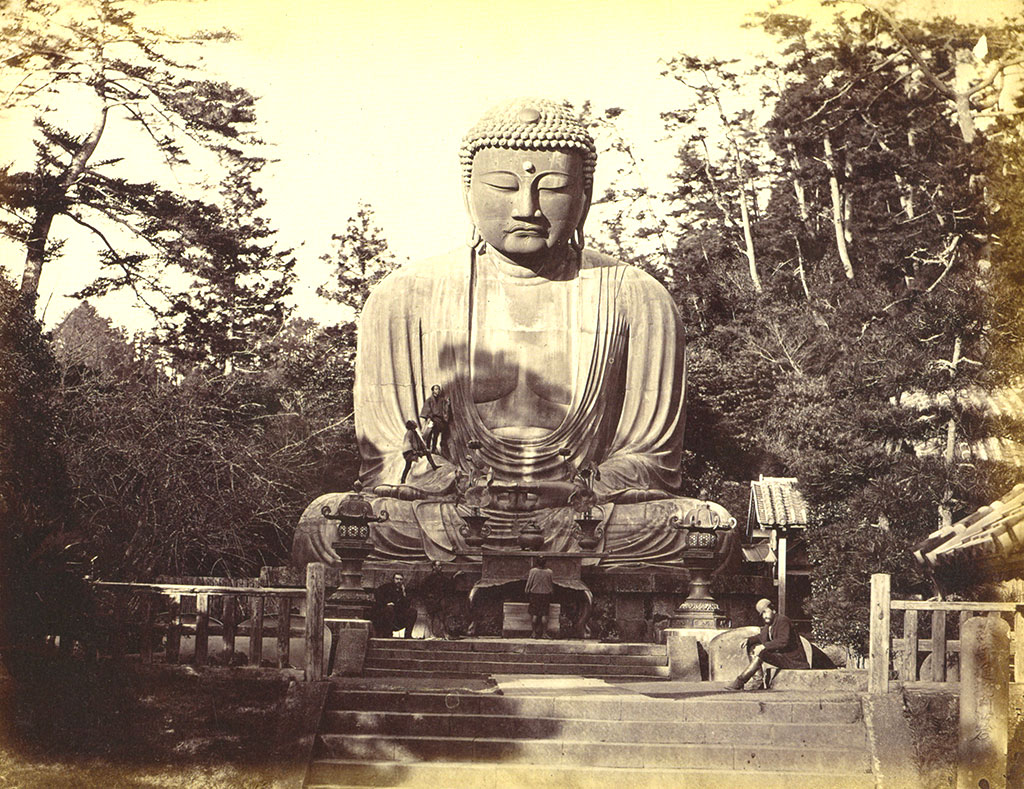 A statue of the Buddha, trees in the background, two people standing on the statue's right forearm