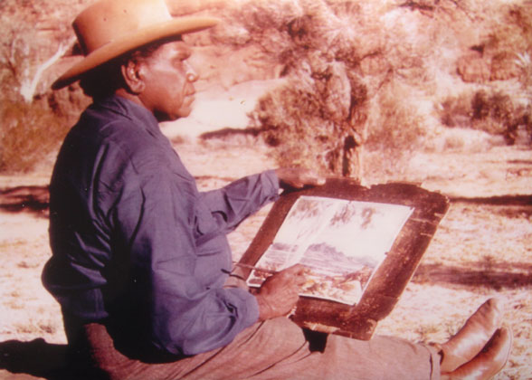 Man, seated on the ground, painting on a piece of paper on a board held on his lap