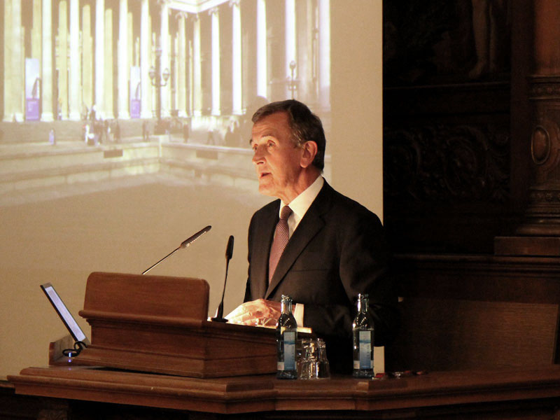 Neil MacGregor shown from the waist up, standing at a lectern. On a screen to his right, a partial image of of a building with columns is projected.