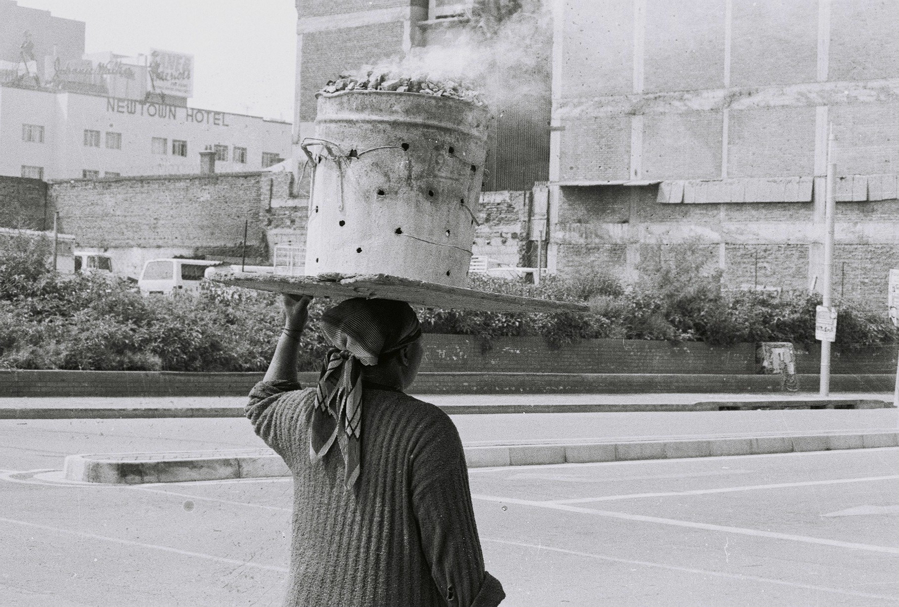 black and white photograph of a woman carrying a bucket of hot coals on her head, seen from the back