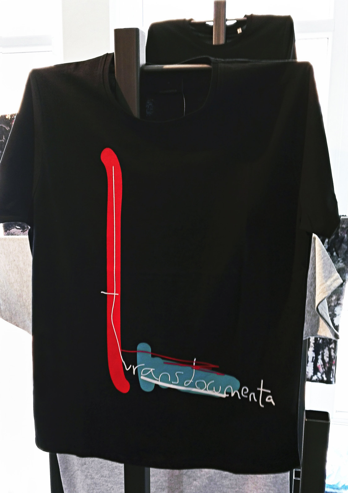 Black t-shirt with transdocumenta logo, hanging on a rack